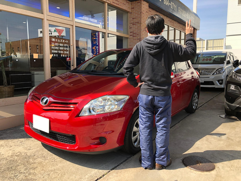 納車された赤いトヨタオーリスの前で記念撮影をするお客様を後ろから撮影した写真