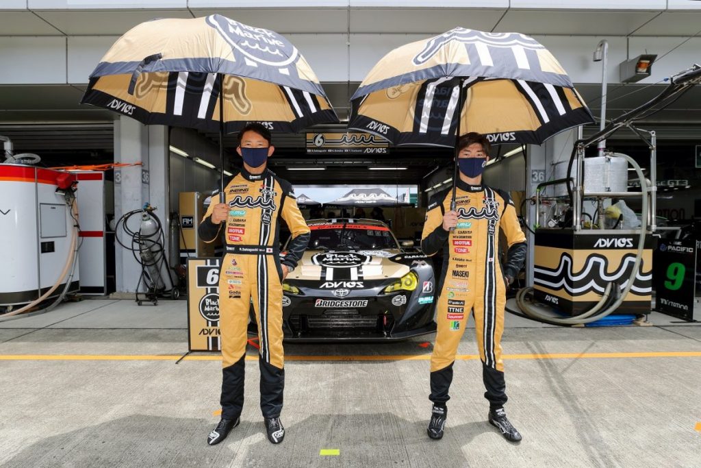 黄色いレーシングスーツを着た2名のドライバーがレースカーの前で傘を差して立つ写真