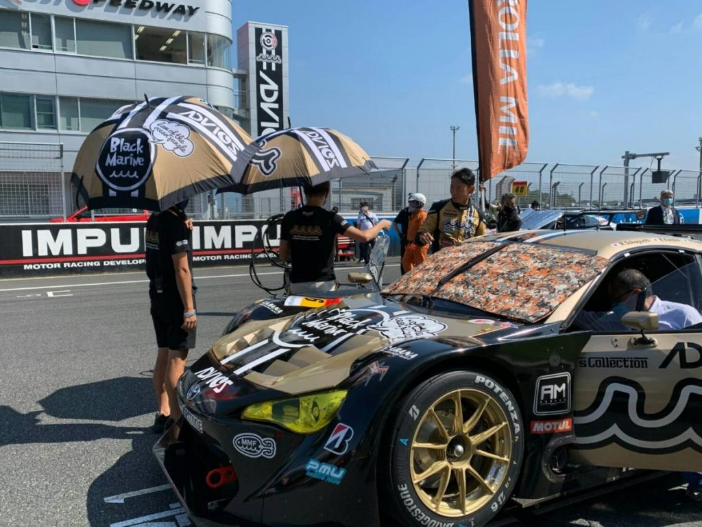 パラソルを差すスタッフとドライバーが並ぶピットエリアでレースカーを撮影した写真