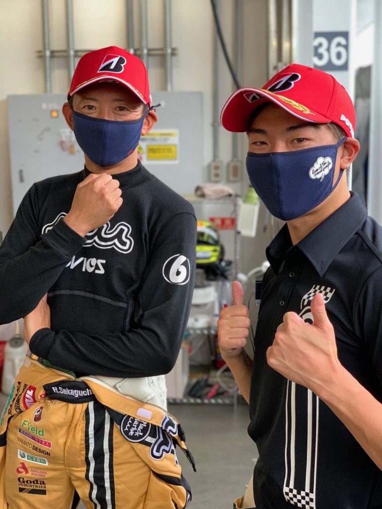 レーシングスーツ姿のドライバーとチームメンバーがピットでポーズをとる写真