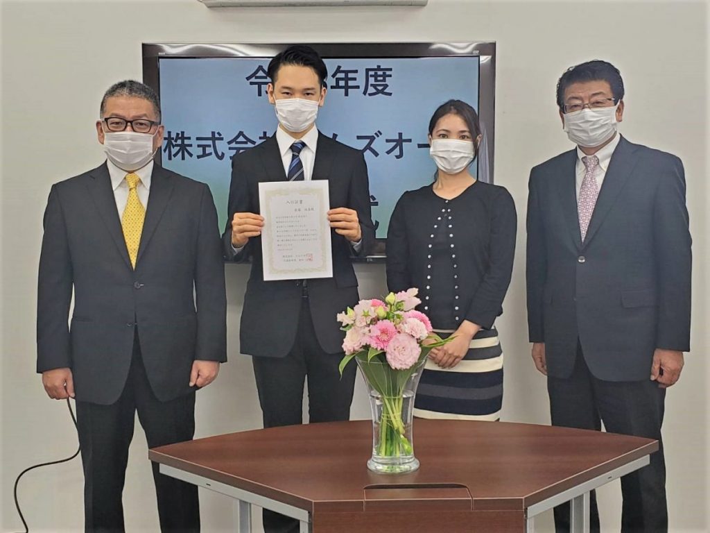 入社式で辞令書を手にした新入社員を囲む社員たちの記念写真
